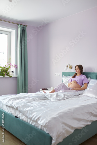 A pregnant woman sits in bed of her domestic bedroom and looking oout the window. Female enjoying her pregnancy state. photo