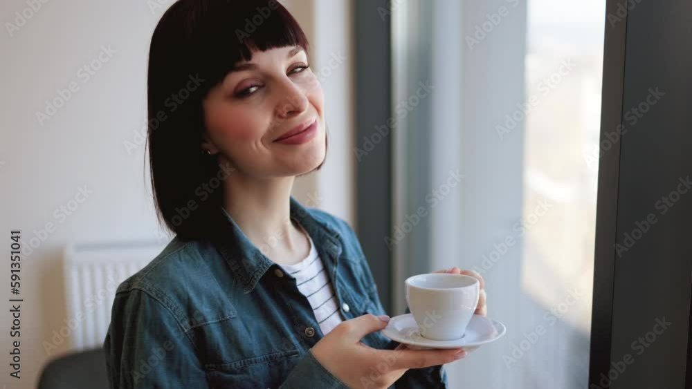Beautiful dark haired lady in denim shirt enjoying aroma coffee during free time at modern office. Caucasian young woman standing with cup in hands and looking at panoramic window. Beautiful dark haired lady in denim shirt enjoying aroma coffee during free time at modern office. Caucasian young woman standing with cup in hands and looking at panoramic window.
