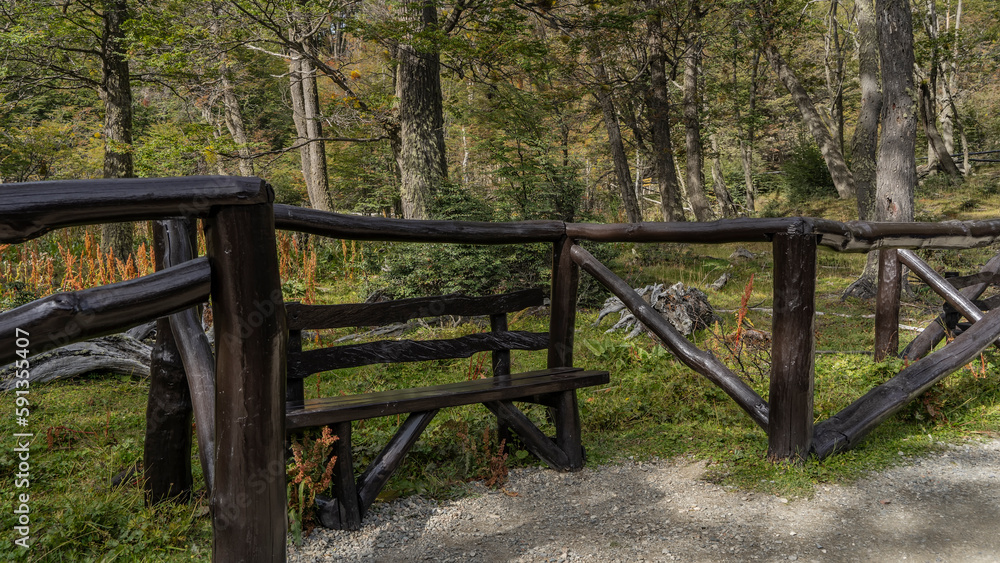 A picturesque wooden bench stands on the side of the road. There is a log fence nearby. Behind- the forest. Tierra del Fuego National Park. Argentina A picturesque wooden bench stands on the side of the road. There is a log fence nearby. Behind- the forest. Tierra del Fuego National Park. Argentina