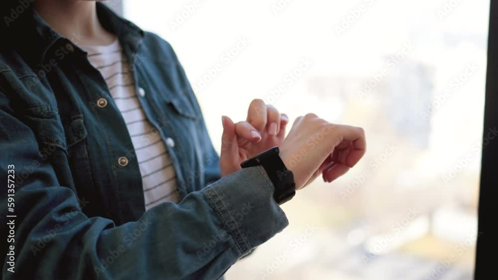 Crop of female office worker touching smartwatch on wrist while standing at workplace near window. Modern wireless gadget for work. Concept of electronic accessories. Crop of female office worker touching smartwatch on wrist while standing at workplace near window. Modern wireless gadget for work. Concept of electronic accessories.
