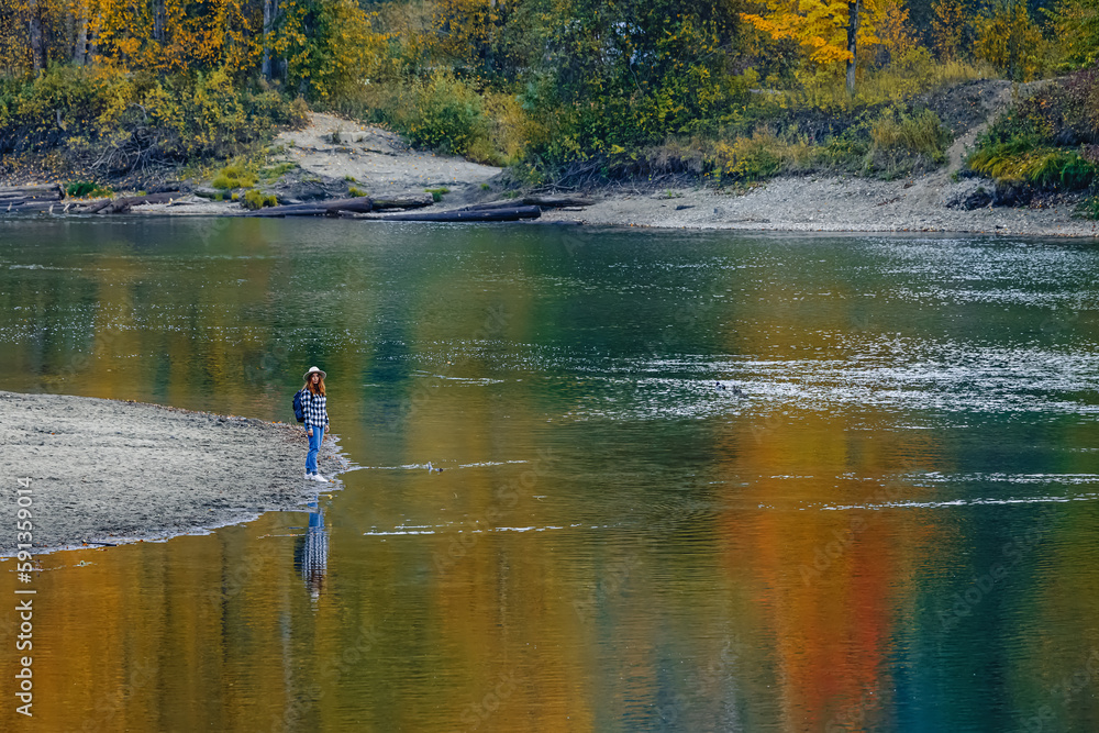 Woman standing on the bank of calm river or lake with reflection of fall thees in the water. Autumn landscape.