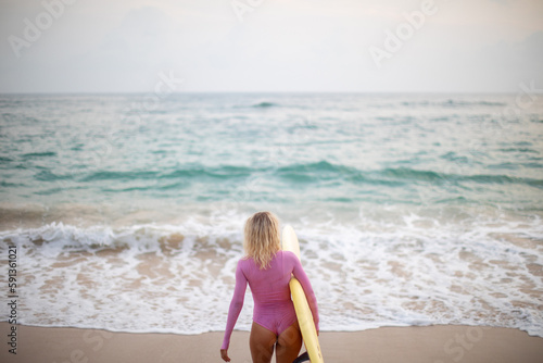 A woman surfer in a pink swimsuit and with a yellow surfboard enters the ocean.