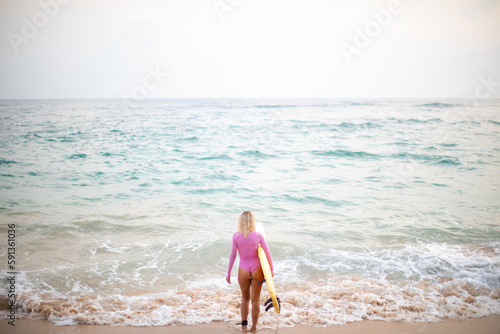A woman surfer in a pink swimsuit and with a yellow surfboard enters the ocean.