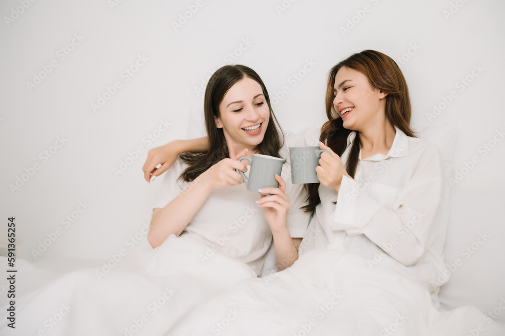 Smiling young sisters lovely couple sitting on white bed together with tablet, smartphone and laptop in bedroom. Homosexual women or Lesbian concept.. Smiling young sisters lovely couple sitting on white bed together with tablet, smartphone and laptop in bedroom. Homosexual women or Lesbian concept..