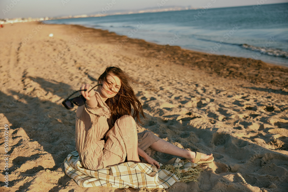 a woman in dark sunglasses sits near the sea on a blanket in windy weather