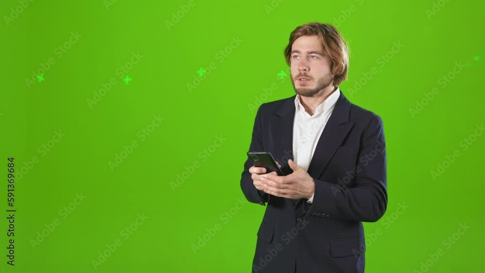 Side view of angry, irritated man standing in studio, holding, using smartphone, typing. Handsome businessman wearing costume and shirt. Isolated on green studio background. Side view of angry, irritated man standing in studio, holding, using smartphone, typing. Handsome businessman wearing costume and shirt. Isolated on green studio background.