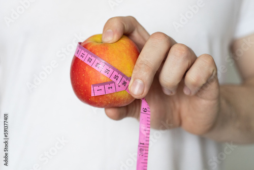 A man in a white T-shirt holds in his hand a red apple with a measuring ribbon close-up. The concept of proper nutrition and healthy lifestyle