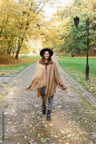 brown-haired woman in a black hat and poncho walks in  autumn park