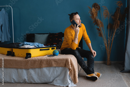 Attractive traveler man with suitcase sit on cozy comfort bed inside modern apartment room speak on cellular, making hotel reservation on phone from home #591383849