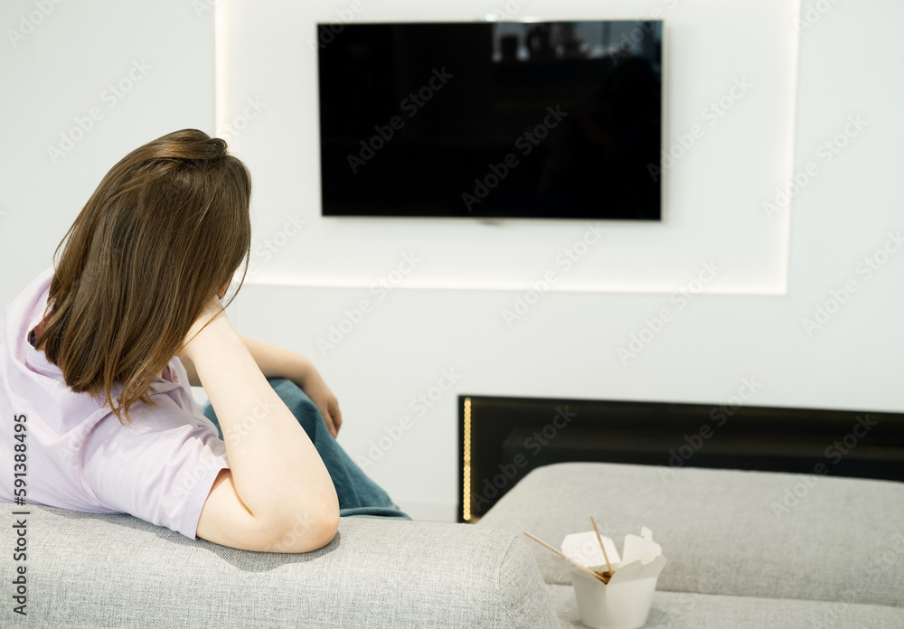 A woman is sitting on a sofa, watching TV, Asian food in a box is nearby, a lonely evening. A woman is sitting on a sofa, watching TV, Asian food in a box is nearby, a lonely evening.