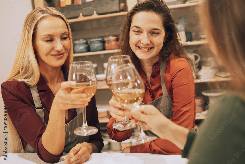 Three cheerful young women friends are having fun in a ceramic workshop. They drink wine and joke, have fun.