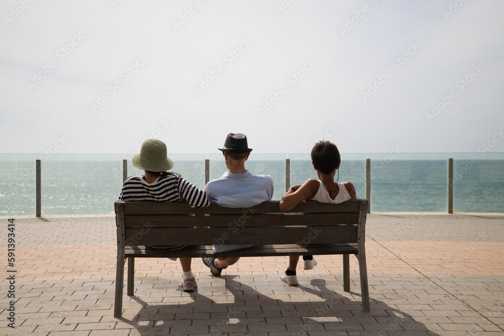 Three people sitting on a bench on the promenade look at the horizon of the Atlantic Ocean, photo taken from behind. People are on a trip in Cadiz, Spain. Concept travel and vacation.
