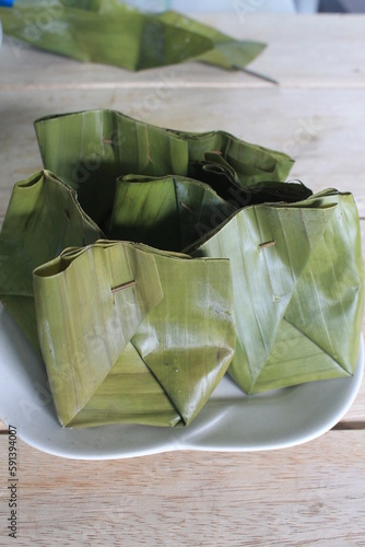 A traditional Indonesian food called sarang gesing made of flour, sugar and banana wrapped in banana leaves inside a white plate on a wooden table

 photo