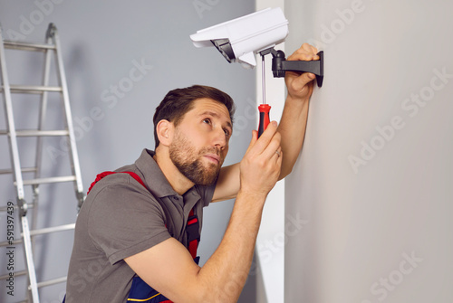 Male service worker installing a security camera in a house or apartment building. Young man in a uniform sets up a new, modern, electronic CCTV surveillance video camera on a white wall at home
