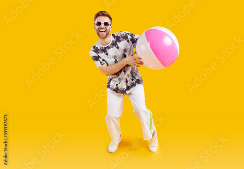 Smiling happy guy in sunglasses holding inflatable sea ball. Full length portrait of joyful young man wearing summer casual clothes standing on isolated yellow studio background