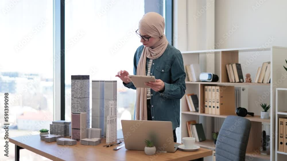 Smiling arabian lady in beige hijab working on digital tablet with open laptop placed near architectural models on office desk. Design professional developing construction project using technologies. Smiling arabian lady in beige hijab working on digital tablet with open laptop placed near architectural models on office desk. Design professional developing construction project using technologies.