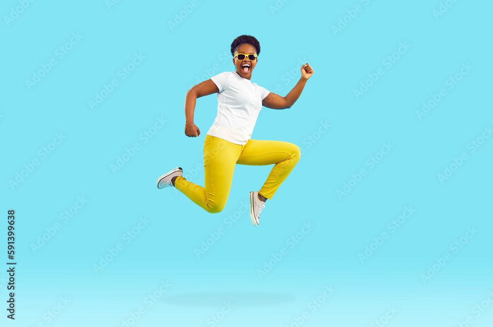 Full body length side profile view portrait happy excited Afro American woman in white tee, yellow pants and cool sunglasses jumping, running and flying high up in air on bright blue studio background