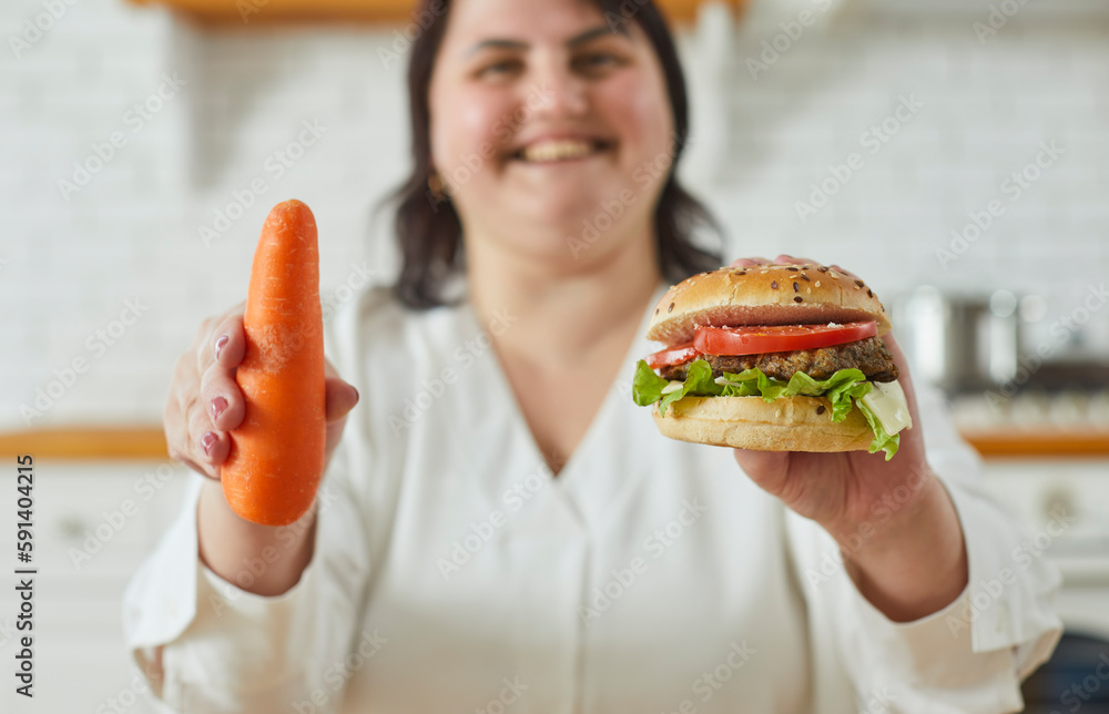 Young overweight plus size fat woman standing in kitchen at home and holding burger in one hand and carrot in other thinking what to choose. Diet restrictions, losing weight concept. Selective focus Young overweight plus size fat woman standing in kitchen at home and holding burger in one hand and carrot in other thinking what to choose. Diet restrictions, losing weight concept. Selective focus