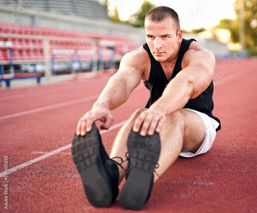 Young muscular athletic runner man stretching and touching his feet on a running court in sitting position before starting of running - Jogging and flexibility concept - Focus on the runner's shoe