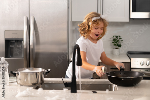 Excited child boy washing the dishes in the kitchen sink. Child with sponge with dish washing liquid is doing the dishes at home kitchen. Cleaning dishware kitchen sink sponge washing dish.