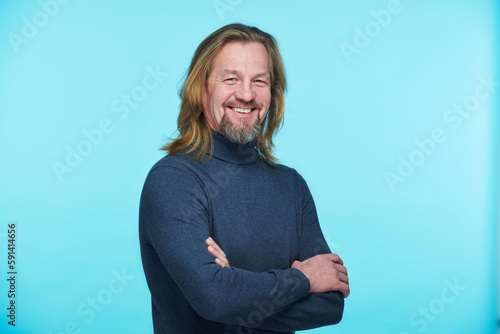 Portrait of mature man with long hair smiling at camera standing with his arms crossed against blue background