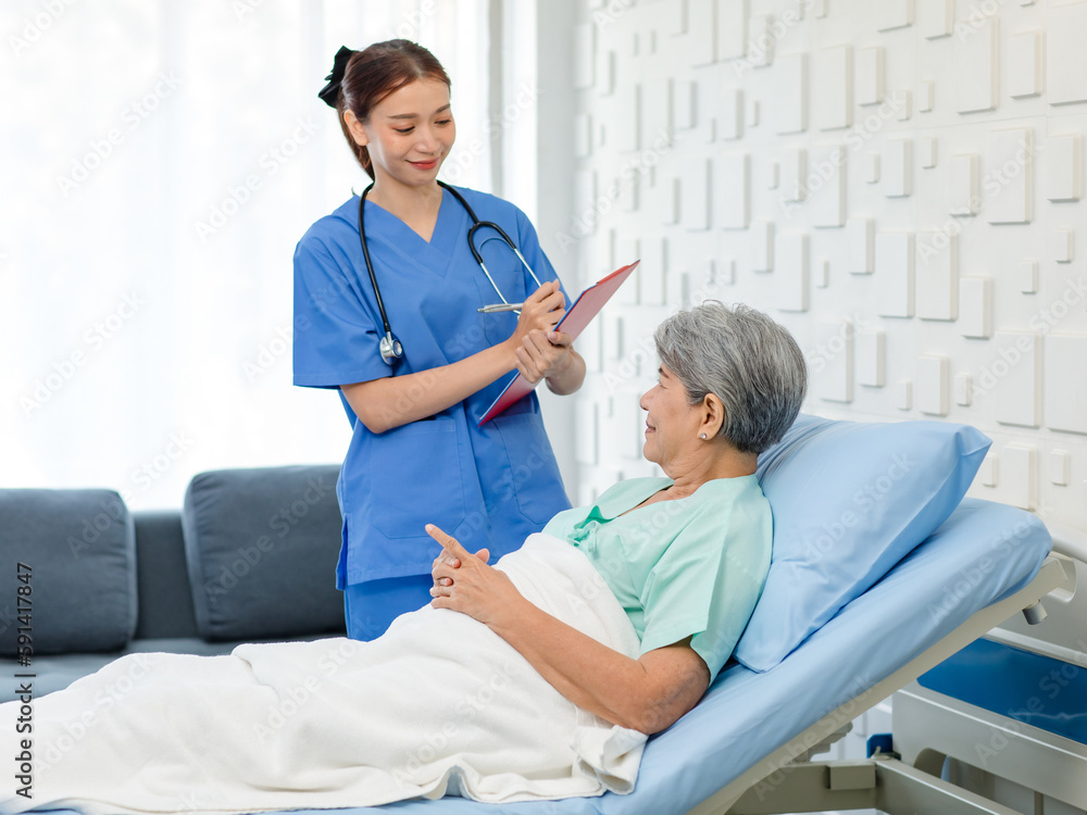 Fototapeta premium Asian professional successful female internship nurse in blue uniform with stethoscope visiting old senior elderly pensioner woman patient laying lying down on bed writing symptoms down on clipboard