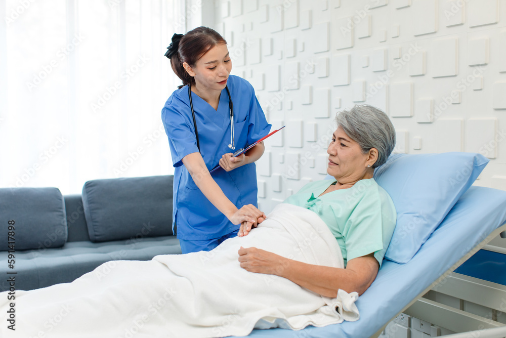 Obraz premium Asian professional successful female internship nurse in blue uniform with stethoscope visiting old senior elderly pensioner woman patient laying lying down on bed writing symptoms down on clipboard