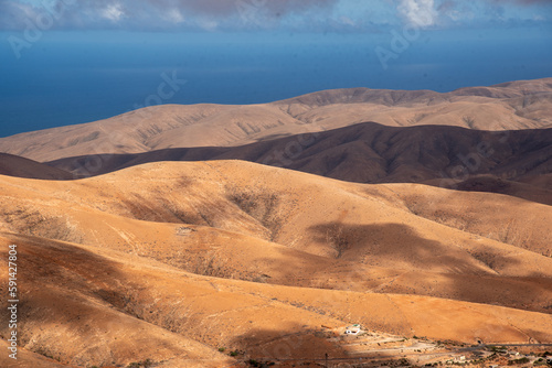  Vista panor  mica de un impresionante paisaje volc  nico y des  rtico  con grandes monta  as rocosas y una peque  a Casa Blanca en una esquina  iluminada por la luz del sol en Fuerteventura  Islas Canaria