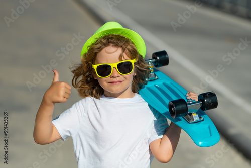 Child boy holding longboard on street. Kid with pennyboard outdoor. Outdoor portrait of cheerful little kid with penny board. Excited child with thumb up, street portrait close up.