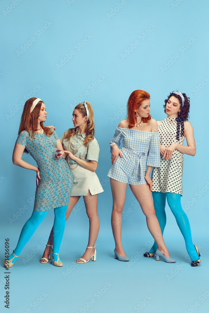 Fototapeta premium Friends meeting. Full-length portrait of attractive young women posing in stylish clothes against blue studio background. Concept of retro style, fashion, beauty, elegance, 60s, youth. Pin-up style