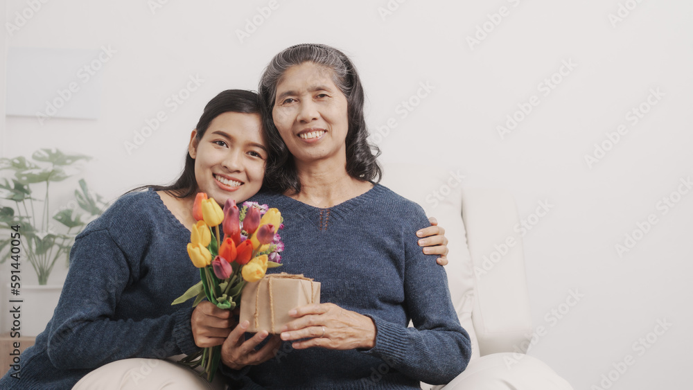 Gift, present, flowers, Mother's day concept, young adult female daughter congratulate excited asian elderly mother at sofa with birthday anniversary, two generations family photo, real people. Gift, present, flowers, Mother's day concept, young adult female daughter congratulate excited asian elderly mother at sofa with birthday anniversary, two generations family photo, real people.