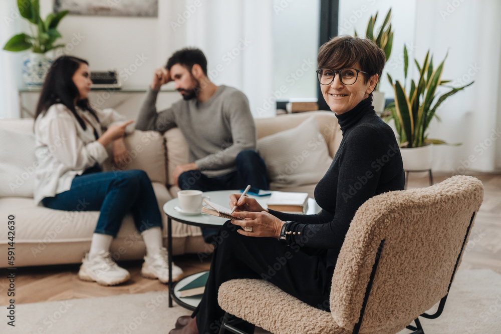 Senior woman psychologist smiling at camera and writing down notes during therapy session with married couple Senior woman psychologist smiling at camera and writing down notes during therapy session with married couple