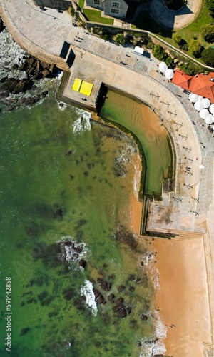 Aerial view of a coastal building with a pool on a sandy beach with green water on a sunny day