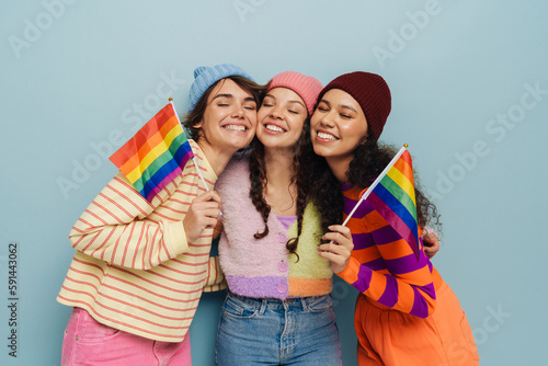 Three cheery girls holding rainbow LGBT flags and hugging isolated over blue background