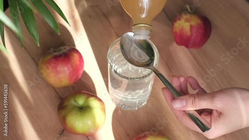 woman carefully pours apple cider vinegar into a spoon, adding it to a glass of water to create a healthy, invigorating drink. Drinking Apple Cider Vinegar: From Weight Loss to Improved Digestion photo