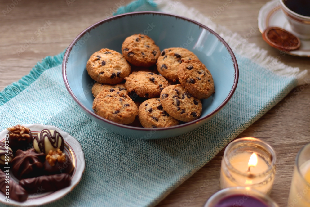 Books, reading glasses, e-reader, plate of chocolate pralines, bowl of cookies, cups of tea and lit candles on the table. Selective focus. Books, reading glasses, e-reader, plate of chocolate pralines, bowl of cookies, cups of tea and lit candles on the table. Selective focus.