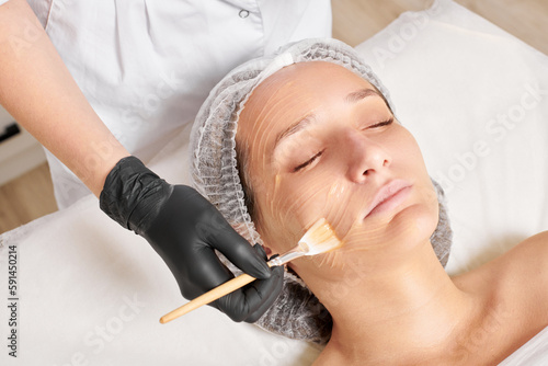 Beautician applies honey mask on woman face for moisturize face skin, anti aging cosmetic procedure in beauty spa salon. Cosmetologist in black gloves holds cosmetic brush for applying honey mask photo