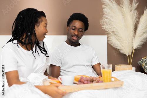 Happy delighted African Americann black couple lying in bed holding tray with tasty breakfast smiling at camera happy morning at weekends. photo