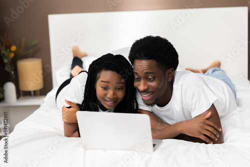 Curious surprised dark skinned couple lying on bed in front of laptop smiling eatching comedy movie film. Happy family concept.