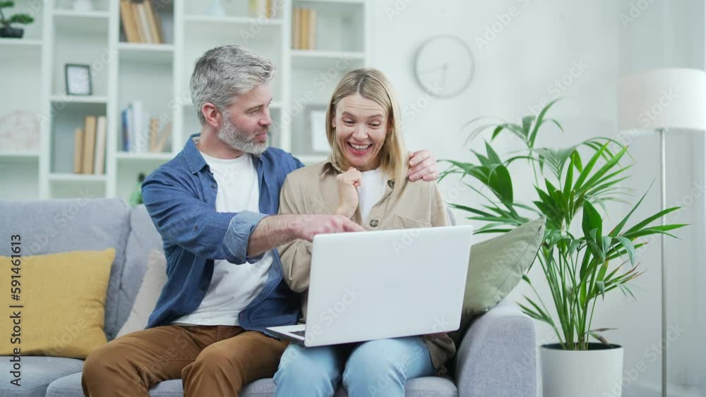 Happy married couple doing online shopping using laptop while sitting on sofa in room at home. Mature smiling wife and husband choose a product in an online store, satisfied with a good promotion