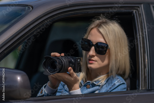 Young blonde woman in sunglasses takes pictures on a professional camera while sitting in a car. Concept of journalism, detective, papparation