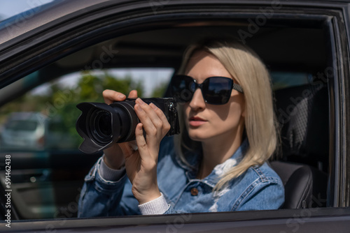 Woman in sunglasses with a camera sits in a car and takes pictures with a professional camera, a private detective or a paparazzi spy. Journalist is looking for sensations and follows celebrities.