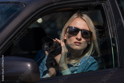 Woman in sunglasses with a camera sits in a car and takes pictures with a professional camera, a private detective or a paparazzi spy. Journalist is looking for sensations and follows celebrities.