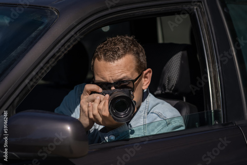 Man in sunglasses with camera sits inside car and takes pictures with professional camera, private detective or paparazzi spy. Journalist seeks sensation and follows celebrities. © Volha
