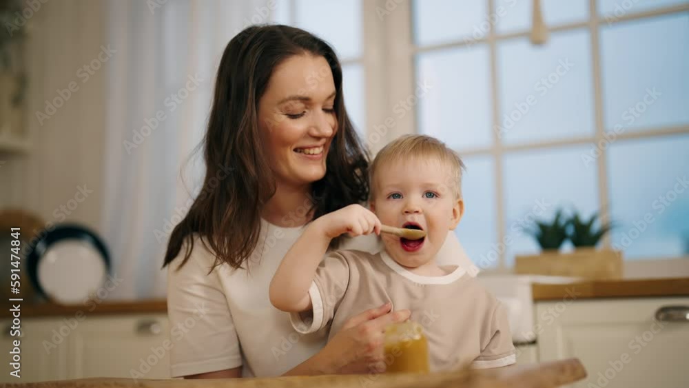 Happy smiling positive mom looks on baby girl eats herself fruit or vegetable puree from jar with spoon sits on kitchen at home. Babyhood, childhood, maternity leave, rising child motherhood concept. Happy smiling positive mom looks on baby girl eats herself fruit or vegetable puree from jar with spoon sits on kitchen at home. Babyhood, childhood, maternity leave, rising child motherhood concept.