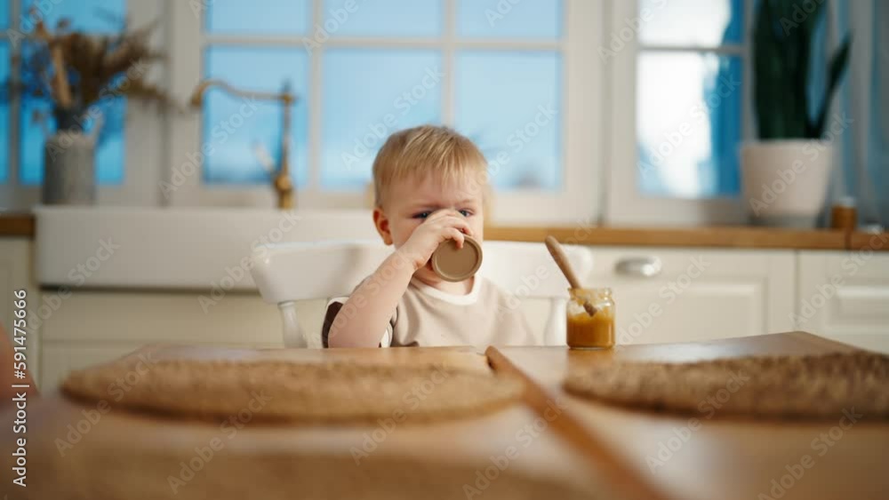 Funny toddler baby girl eats fruit or vegetable puree from lid of jar sits at table in kitchen at home. Independent child eats by herself looking at camera. Childhood, babyhood, adorable kids concept. Funny toddler baby girl eats fruit or vegetable puree from lid of jar sits at table in kitchen at home. Independent child eats by herself looking at camera. Childhood, babyhood, adorable kids concept.