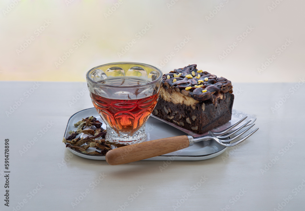 Piece of Homemade Toffee cake layer with cheesecake, is decorated with sliced cashew nuts with butterfly pea flower tea (Clitoria) and dried butterfly pea flowers served with fork on white iron plate.