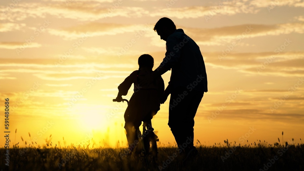 Father helps his daughter ride bike. Father teaching little daughter to ride bike in park. Child rides bike. Child, dad play together, sunset. Chidhood dream. Kid Girl learns to ride bike.Happy family