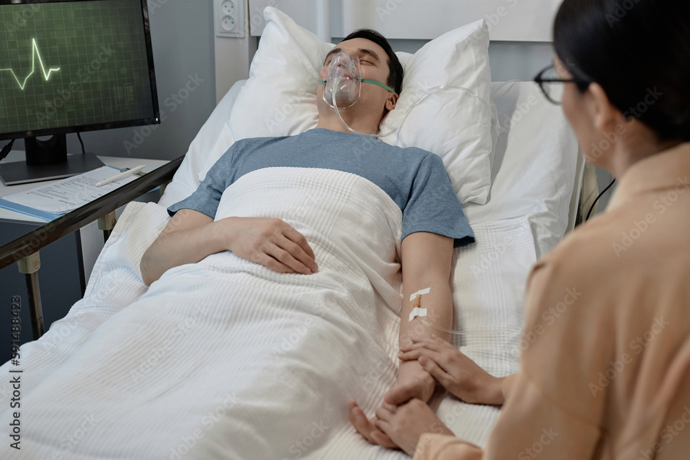 Young woman visiting her sick husband in hospital ward, she sitting and holding his hand while he lying on bed with oxygen mask