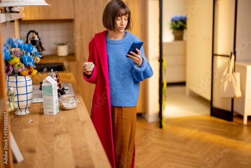 Фотография Woman rushes out of home putting on a coat, finishing her coffee and looking at smart phone
